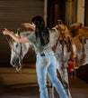 A person wearing jeans and a green shirt adjusts the bridle of a saddled gray horse indoors, highlighting the elegance of an Argentine leather Rambler by Zilker Belts.