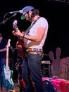 Male musician wearing a light brown leather belt with black and white stitching. He plays the guitar.